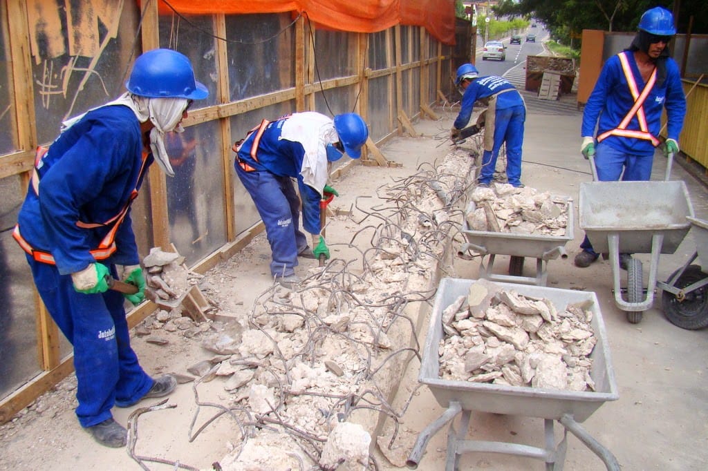 Quatro trabalhadores da construção civil em uniformes azuis e capacetes trabalham em uma obra ao ar livre, quebrando concreto e enchendo carrinhos de mão