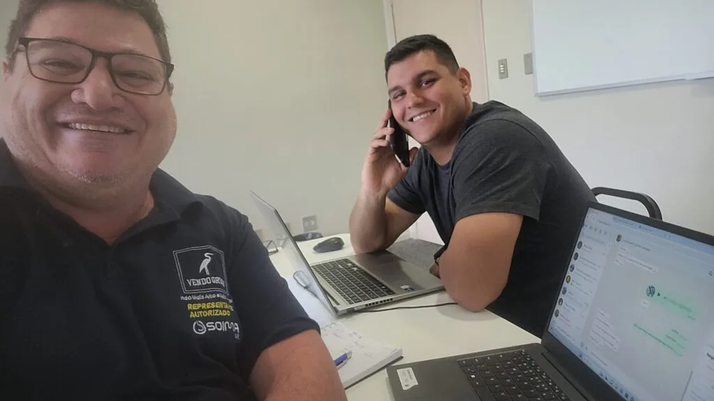 Dois colegas de trabalho em um escritório, um falando ao telefone celular, outro sorrindo para a câmera.