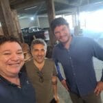 Três homens posam para uma selfie sorrindo em um ambiente rústico com pilares de madeira e uma caminhonete ao fundo.