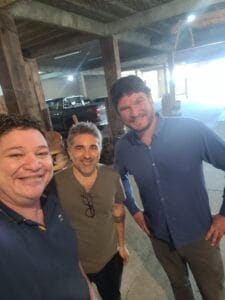 Três homens posam para uma selfie sorrindo em um ambiente rústico com pilares de madeira e uma caminhonete ao fundo.