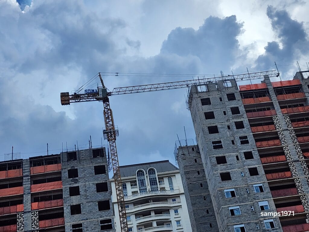 Fotografia de baixo ângulo de um guindaste amarelo no centro, com dois edifícios altos em construção em ambos os lados, mostrando estruturas de concreto e detalhes em laranja, sob um céu nublado com nuvens brancas e cinzas.